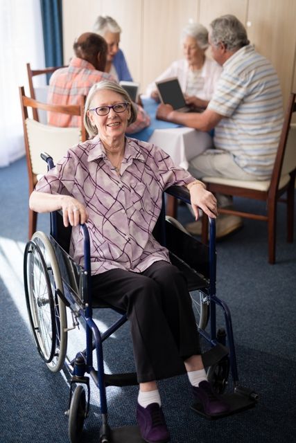 This image shows a smiling senior woman sitting in a wheelchair at a retirement home, with a group of friends socializing in the background. It can be used for promoting elderly care services, retirement homes, health care facilities, and community living for seniors. It highlights themes of independence, support, and social interaction among the elderly.