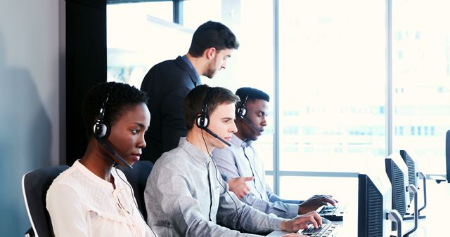 A diverse group of professionals, including African American and Caucasian individuals, are working in a modern call center environment, with copy space. They are equipped with headsets and computers, focusing intently on providing customer support.