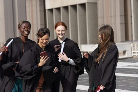Diverse female graduates celebrating on rooftop, checking smartphone and holding diplomas