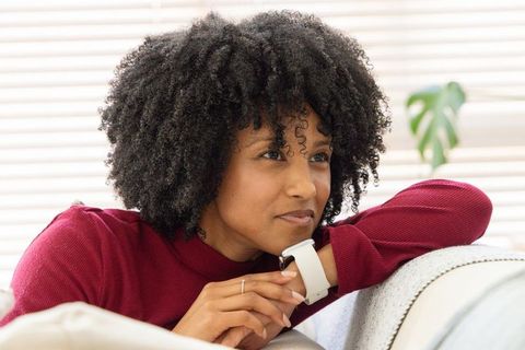 African american woman relaxing on sofa wearing smartwatch, gazing thoughtfully indoors