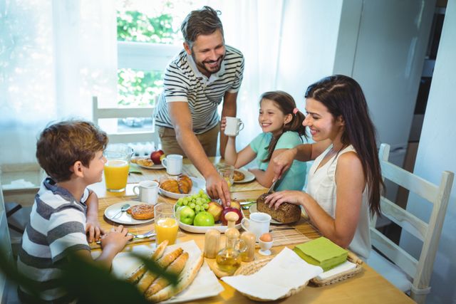 Family of four enjoying breakfast together at home. Parents and children are smiling and bonding over a meal that includes fresh fruits, bread, and juice. Ideal for use in advertisements promoting family values, healthy eating, or breakfast products.