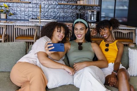 Three friends taking selfie on sofa in modern kitchen-living area, smiling together