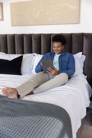 Young man reclining and smiling on bed using tablet in modern minimalist neutral bedroom