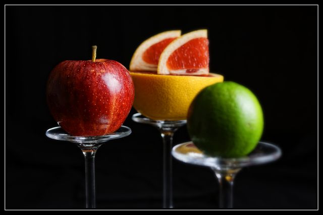 Colorful arrangement of apple, grapefruit, and lime on glass pedestals against dark background emphasizes the vivid colors and textures of the fruits. Ideal for use in marketing materials for health and nutrition, organic food promotions, diet plans, and fresh produce advertisements. The contrasting dark background enhances the vibrancy and appeal of the fresh fruits.