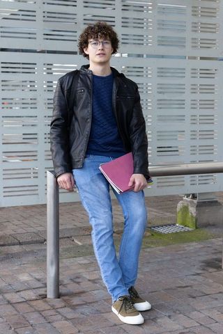 Young man in twenties leaning on metal railing holding maroon folder on urban campus