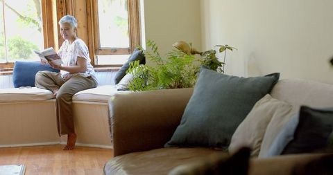 Senior Woman Reading by Window with Comfortable Couch at Home
