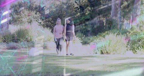 Two women walking in sunlit park wearing athletic wear holding bottle with neon overlay