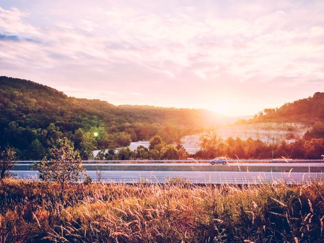Car driving on highway during sunset with rolling hills in background can be used for travel blogs, promotional travel brochures, or road trip advertisements emphasizing tranquility and beauty of nature.