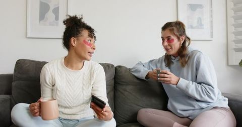 Diverse female friends sitting on couch holding mugs and wearing under-eye gel patches