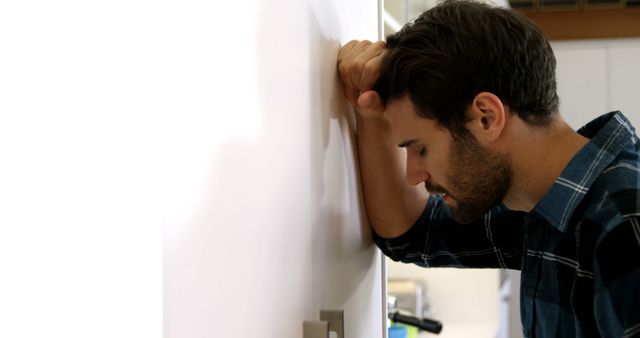 This visual depicts a young man leaning his head against a wall, showing signs of stress, frustration, or worry. He wears a plaid shirt and appears deep in thought or overwhelmed. This visual can be used to illustrate concepts related to mental health, emotional exhaustion, stress management, and coping strategies in various contexts such as articles, therapy websites, and social media posts.