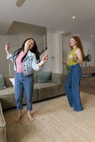 Diverse female friends dancing barefoot in cozy modern living room wearing denim and lime