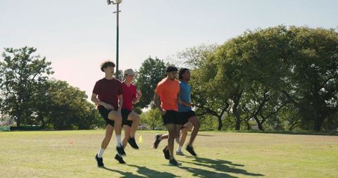 Multiracial teen athletes warming up with high-knee drills on sunny park field
