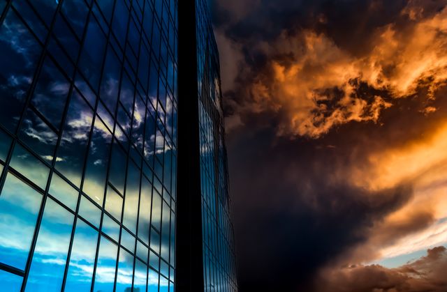 Modern glass building reflecting a dramatic sunset sky with vibrant orange clouds. Ideal for use in real estate marketing, architectural presentations, cityscape imagery, and environmental contrast themes.