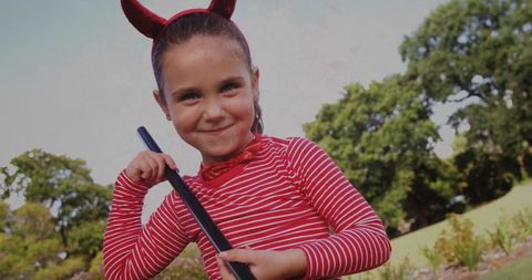 Girl in devil costume holding broomstick smiling in park