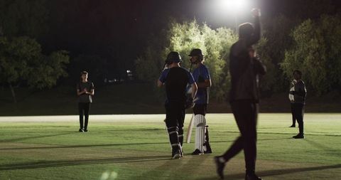 Two batters conferring under floodlights on night cricket pitch, players preparing to bat