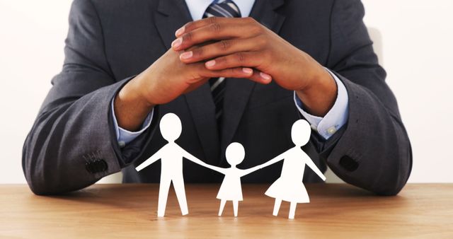 African American businessman in a suit protects a paper cut-out of a family with his hands, symbolizing insurance or security, with copy space. It conveys the concept of family protection and financial planning in a corporate context.