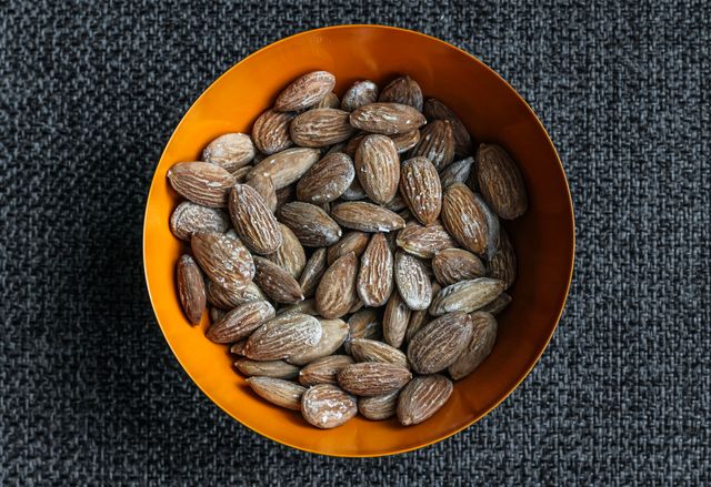 Salted almonds displayed in an orange bowl on dark woven-texture background, perfect for illustrating healthy snack choices, nutrition articles, or recipe blogs. Can be used in food-related promotions and advertisements focusing on healthy eating habits.