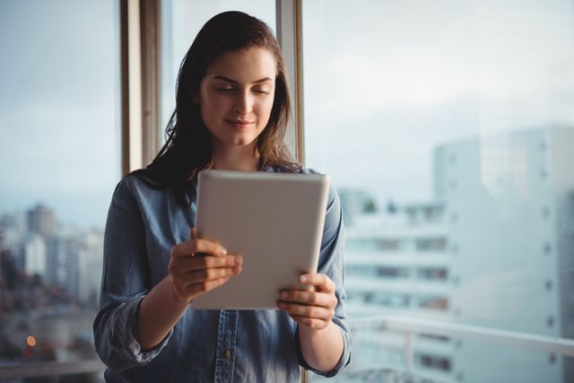Smiling woman holding digital tablet while standing by window at home