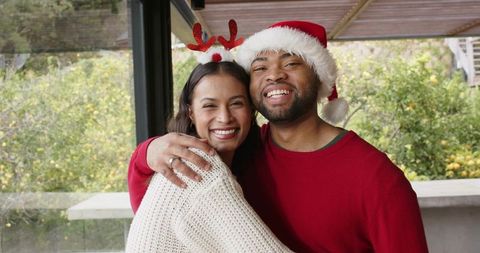 Couple hugging and smiling on patio wearing santa hat and reindeer antlers, celebrating