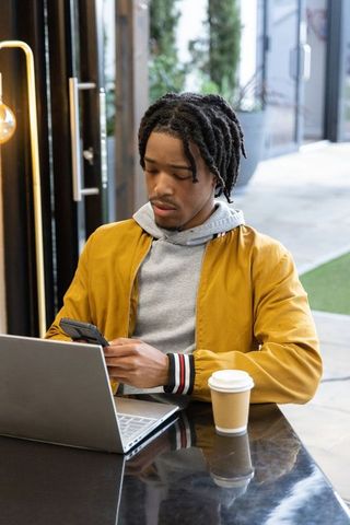 African american man in mustard jacket working on laptop and phone in modern coffeehouse