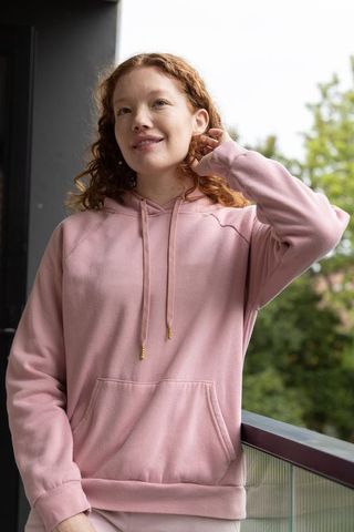 Redhead woman standing on balcony touching curly hair in pink hoodie, relaxed casual