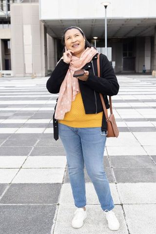 Mature asian woman smiling and talking on smartphone in urban tiled plaza wearing pink scarf