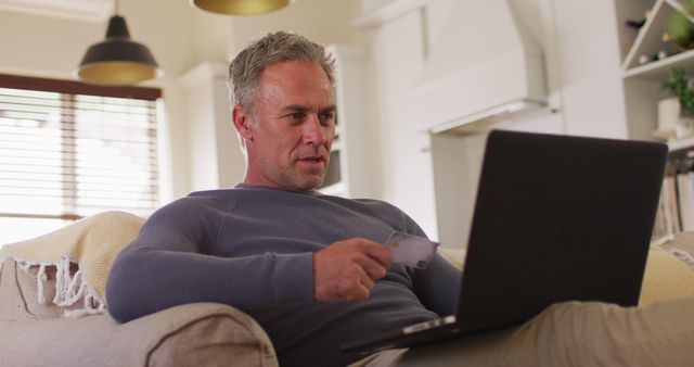 Middle-aged man with grey hair using a credit card while shopping online on a laptop, seated on a couch in a modern, comfortable living room. Can be used for topics related to online shopping, ecommerce, secure payment, remote work, casual lifestyle, technology use at home.