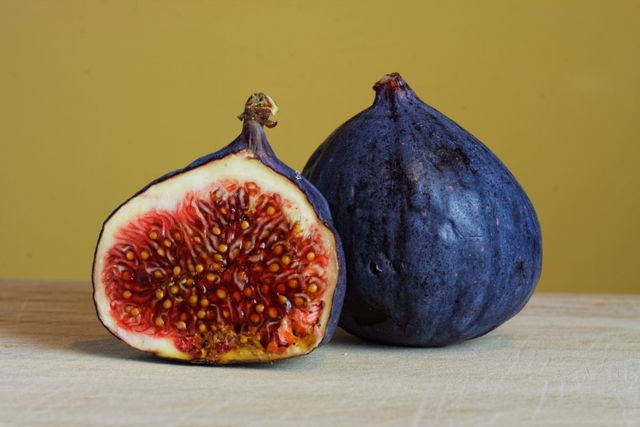 Close-up view of a fresh fig fruit, showcasing one whole fig and a halved fig, both resting on a beige surface against a yellow background. The vibrant colors and intricate details of the fruit seeds are highlighted. Ideal for use in health and lifestyle articles, food and cooking blogs, nutrition websites, and advertising campaigns emphasizing fresh, organic, and healthy produce.