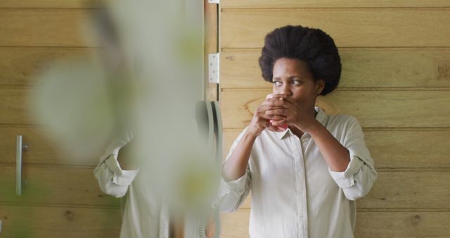This image depicts a woman with afro hair drinking coffee while standing in a modern, interior space featuring a wooden wall. The scene suggests a tranquil and serene moment, making it suitable for use in lifestyle blogs, wellness articles, or advertisements focused on self-care, relaxation, and home living.