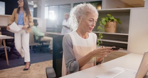 Mature Woman Multitasking at Office Desk with Smartphone and Laptop