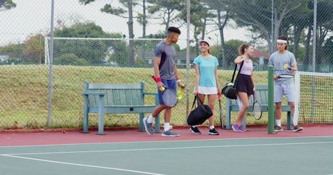 Multiracial young adults walking on tennis court carrying rackets and duffel bags