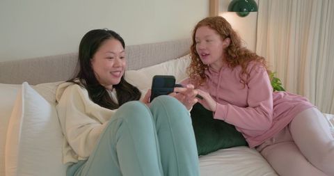 Friends lounging on bed sharing smartphone in cozy pastel loungewear casual home moment