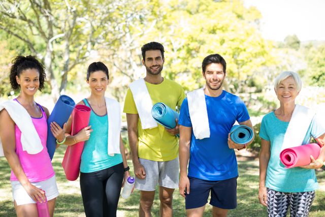 Diverse group standing outdoors in a park, each holding rolled exercise mats and wearing workout clothes with towels on their shoulders. Ideal for promoting outdoor fitness, community exercise events, group fitness classes, wellness programs, and healthy lifestyle activities.