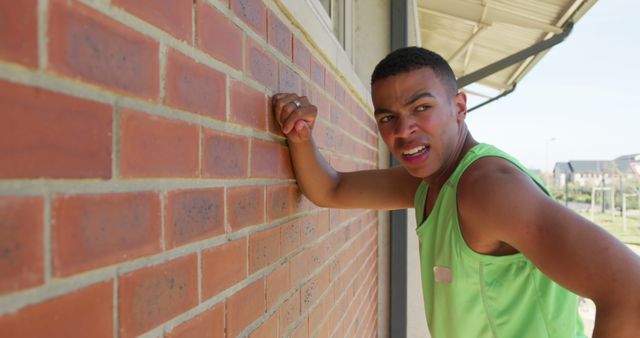 Athletic man in green tank top leaning on a red brick wall outside, possibly showing frustration or exhaustion. Ideal for use in health, fitness, and sports-related content, conveying determination, challenge, or struggle in athletic endeavors.