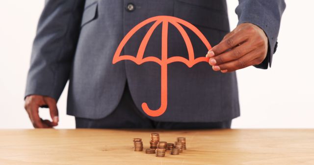 A middle-aged African American businessman is holding an illustration of an orange umbrella over stacks of coins, symbolizing financial protection or insurance, with copy space. It represents the concept of safeguarding investments and the importance of financial security.