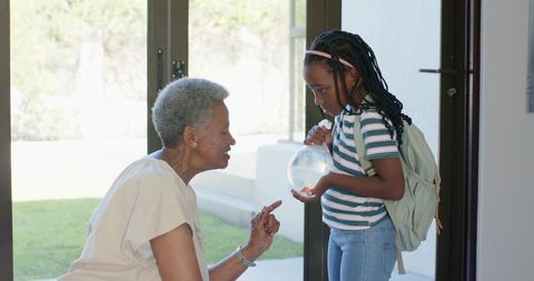 African grandmother kneeling talking to granddaughter holding fishbowl at front door