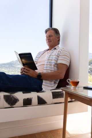 Relaxing senior man reading on sunlit window bench with tea and countryside view