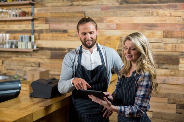 Smiling waiter and waitress using a digital tablet at the counter of a cozy cafe. Ideal for content about modern small businesses, teamwork, customer service, and the use of technology in everyday work settings. Suitable for marketing materials, blog posts about hospitality, and social media content highlighting behind-the-scenes in cafes.