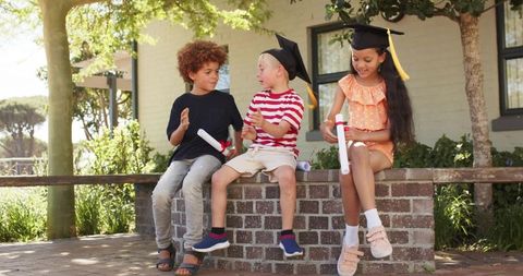 Children celebrating graduation outdoors wearing caps holding diplomas