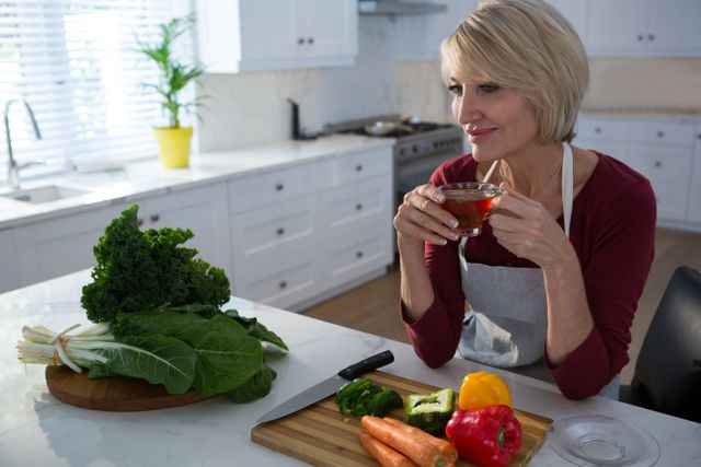 Middle-aged woman sitting at kitchen table, holding cup of lemon tea, surrounded by fresh vegetables. Ideal for use in healthy lifestyle blogs, cooking websites, home decor magazines, and wellness advertisements.