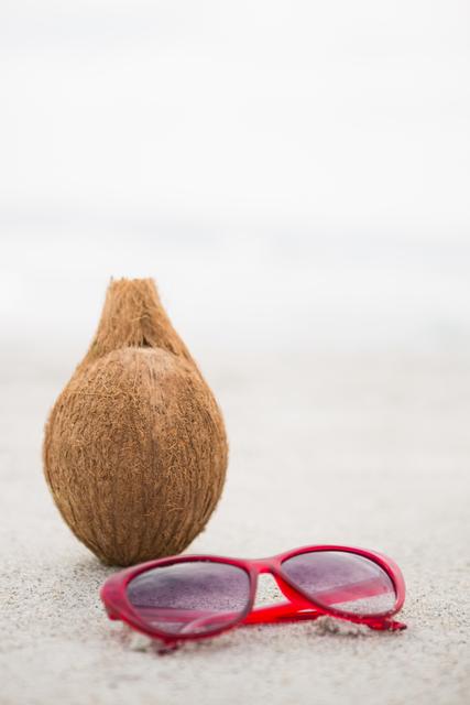Coconut and red sunglasses placed on sandy beach, evoking a sense of tropical vacation and relaxation. Ideal for travel brochures, summer holiday promotions, beach-themed advertisements, and social media posts about tropical getaways.