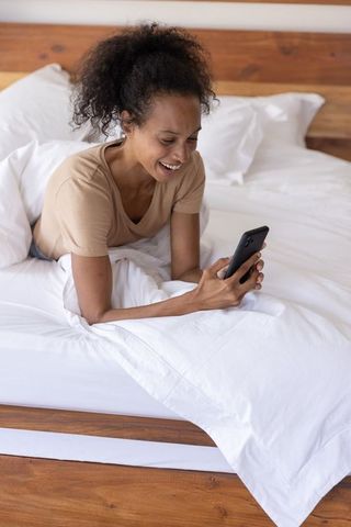 Smiling african american woman lying in bed texting on smartphone enjoying cozy morning