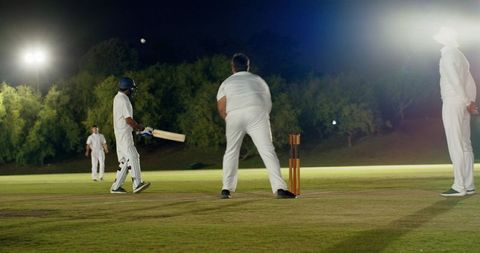 Night cricket match under floodlights, batsman facing bowler near stumps on turf