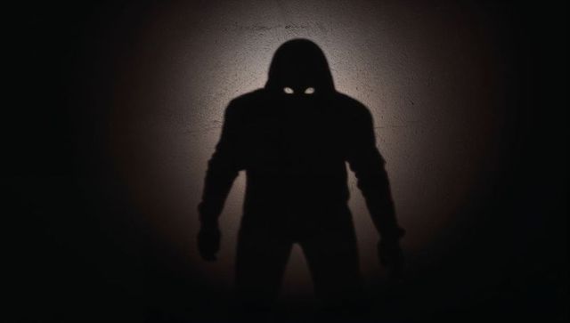 Hooded shadow casting glowing eyes onto textured wall under spotlight