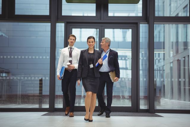 Businesspeople walking together in a modern office environment, holding documents and wearing professional attire. Ideal for use in corporate websites, business presentations, and promotional materials highlighting teamwork, professionalism, and a collaborative work environment.