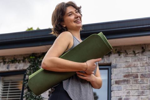 Smiling woman holding green yoga mat on brick patio for casual home workout