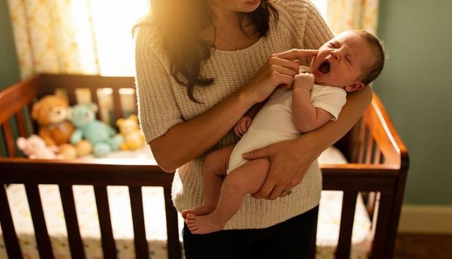 Sunlit mother cradling yawning newborn by wooden crib in cozy nursery with plush toys