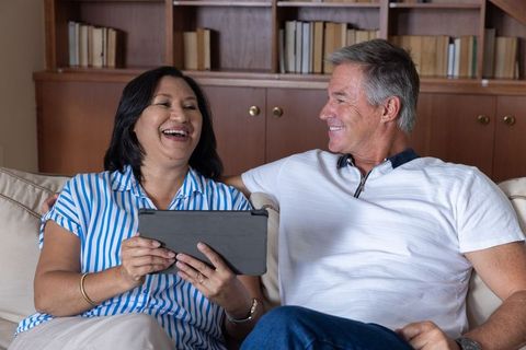 Mature couple laughing and holding tablet while sitting on sofa at home