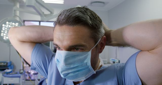 Male doctor in scrubs adjusting his face mask in a hospital room. Ideal for use in healthcare promotions, medical-related articles, safety guidelines, and educational materials focusing on medical professionals and patient care.