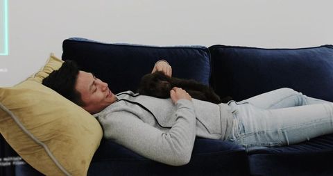 Asian man relaxing on navy sofa stroking puppy in modern cozy living room with neon accent